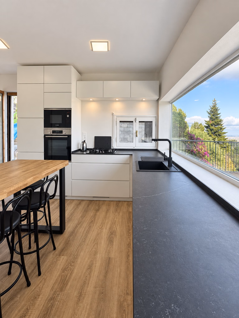 Cocina blanca con encimera oscura, barra de madera y ventana hacia el exterior.